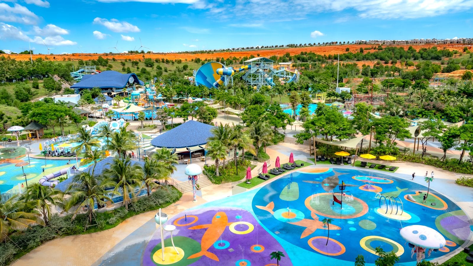A water park laid out among palms — splash pads painted with fish, a wave pool and a looping blue slide — with red sand and a line of wind turbines on the ridge behind