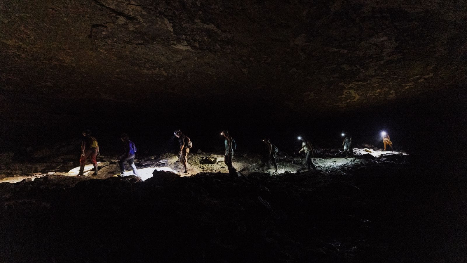 A line of eight people picking their way across a boulder floor deep inside a cave, each lit only by a head torch, the roof lost in the dark above them